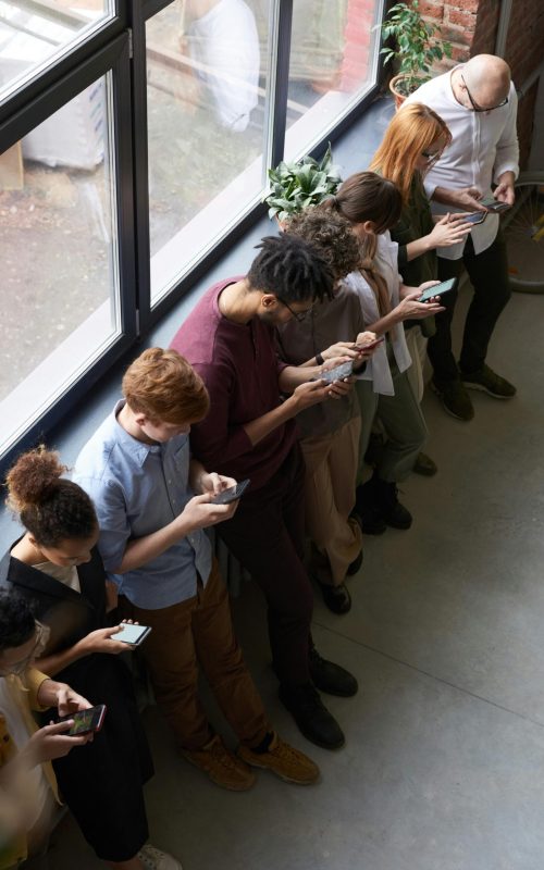 A group of diverse office workers standing by a window using smartphones, capturing modern workplace connectivity.